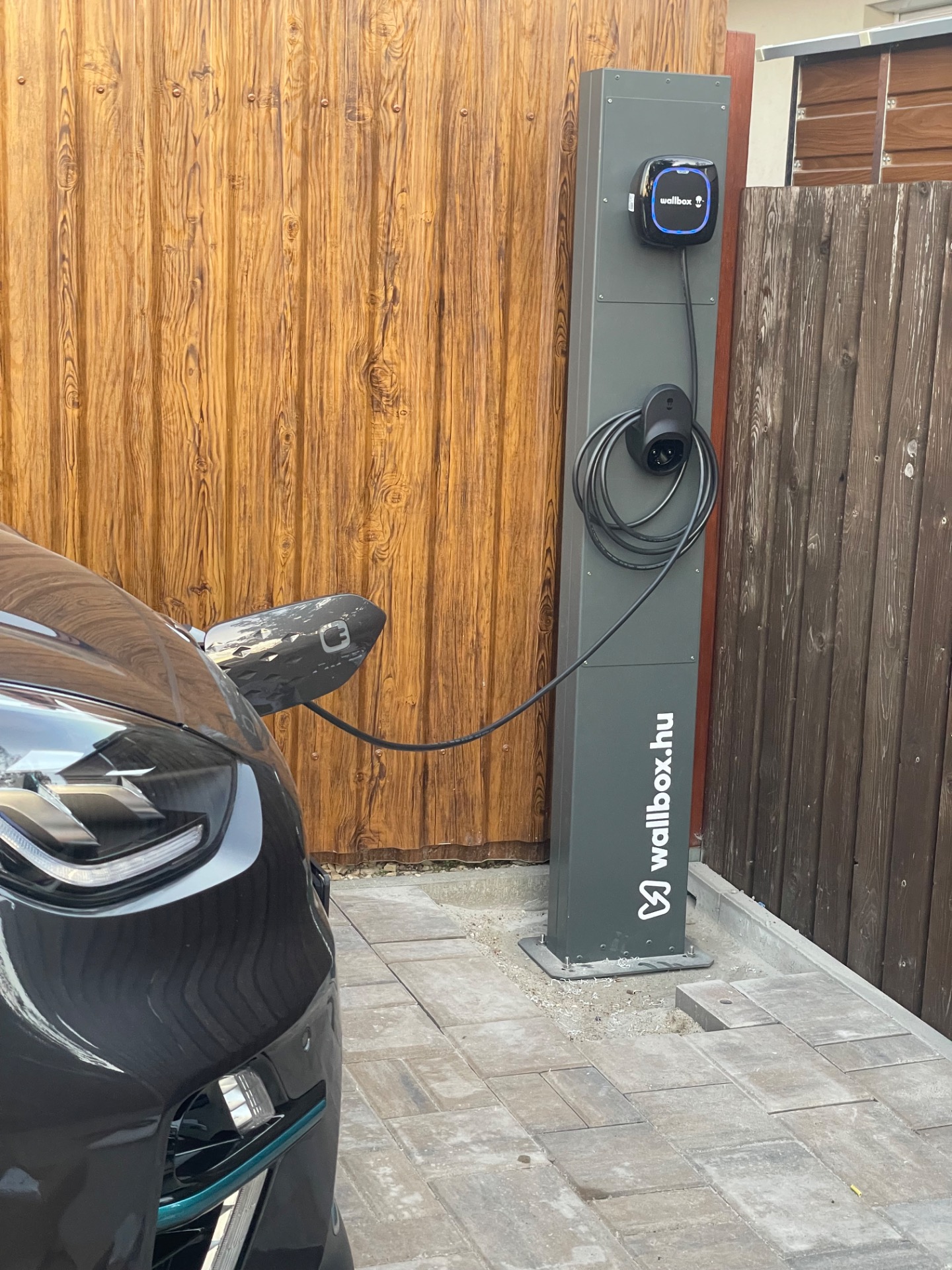 Wallbox töltőoszlop csatlakozó kábellel, fából készült fal mellett, parkoló autó részletével.